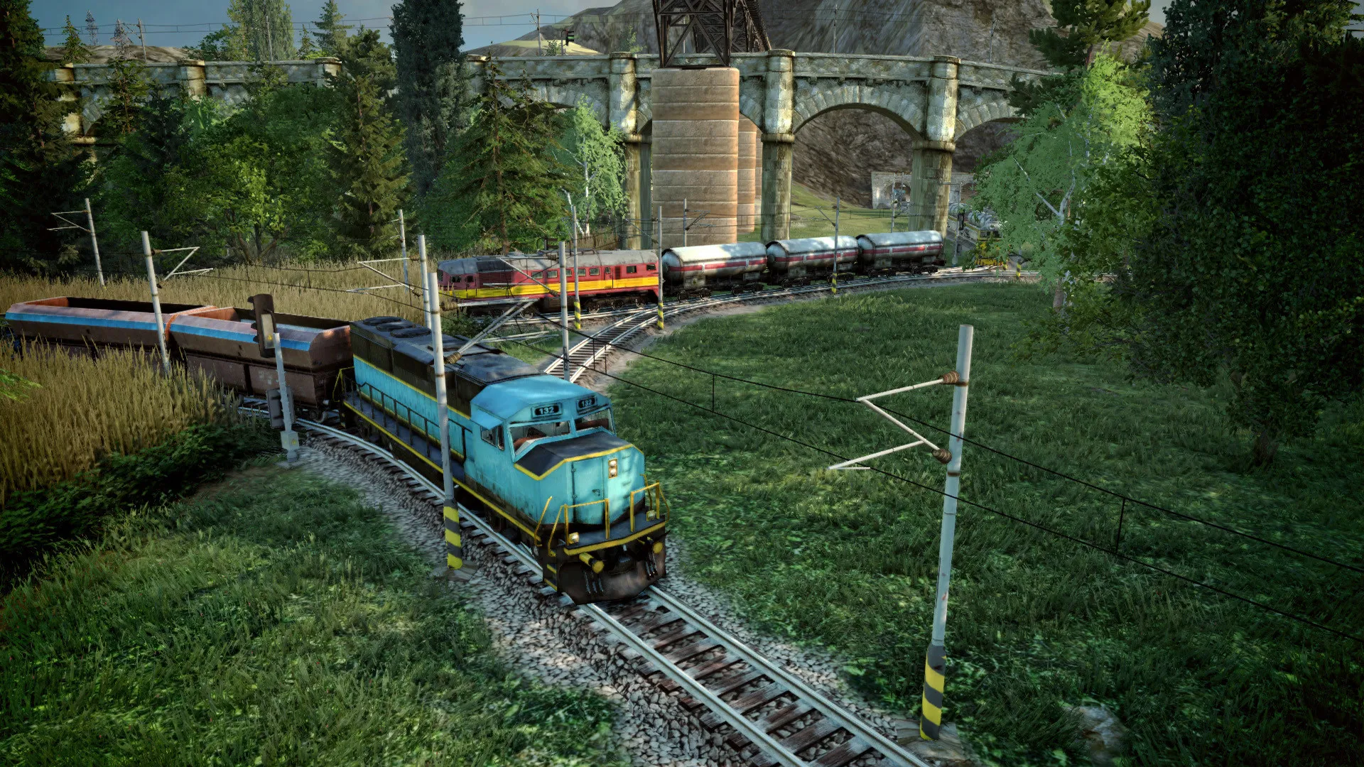 A diesel locomotive hauling freight round a curve through meadows in Mashinky, with a viaduct behind it